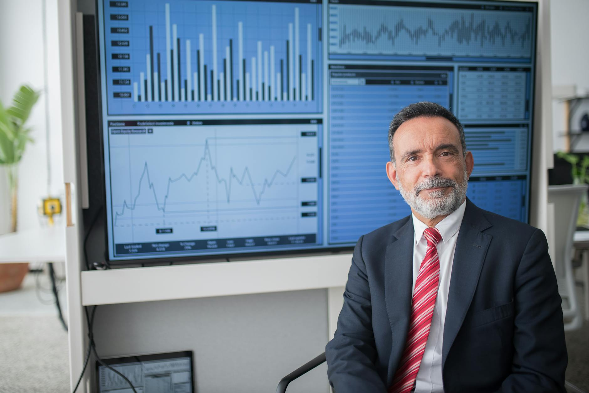 Smiling businessman in a dark suit and red striped tie seated beside a wall of financial charts and graphs on a large screen behind him.